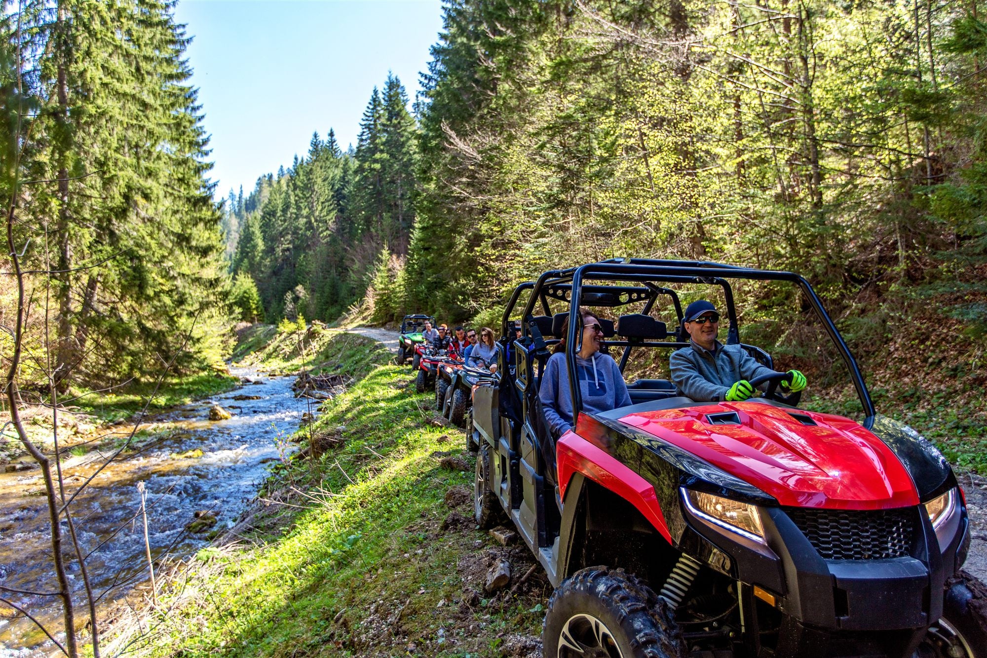 A group of side-by-side rentals rides a wooded trail along a creek.