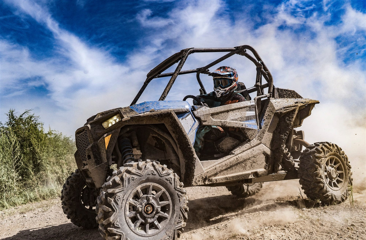 A rider in a helmet drives a mud-covered side-by-side down a dirt trail under a blue sky.
